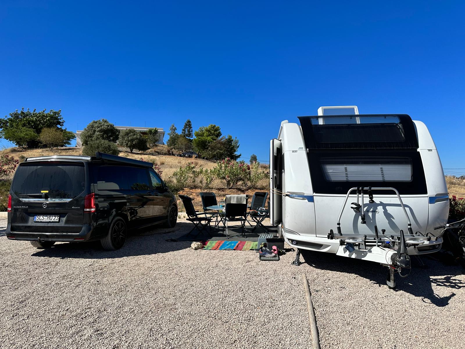 Unser Hobby-Wohnwagen und schwarzes Zugfahrzeug auf einem sonnigen Stellplatz mit Campingstühlen.